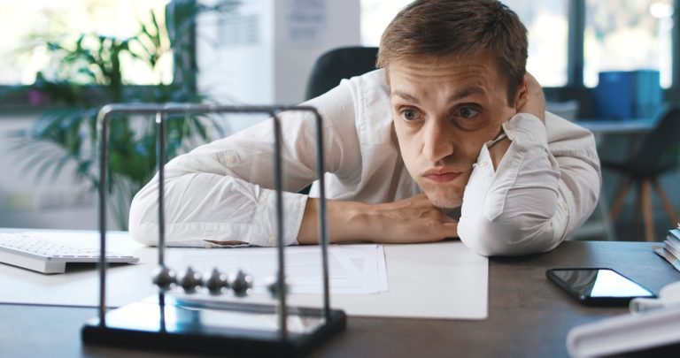 Lazy inefficient businessman sitting at desk and staring at Newton's cradle balls moving